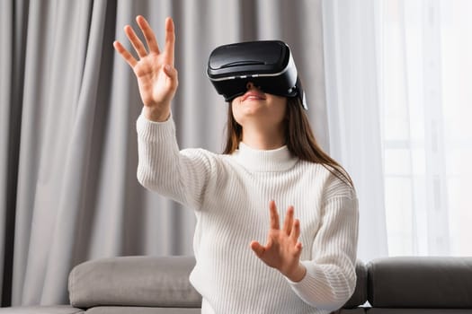 A beautiful caucasian woman uses VR headset touching an invisible screen sitting in the living room.