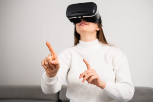 Curious housewife in VR goggles swipes virtual interface to choose game. Young woman rests on couch tasting modern innovations at home