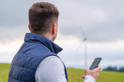 Engineer uses the mobile phone to check the functioning wind turbine. Wind turbine produces green energy against grey sky