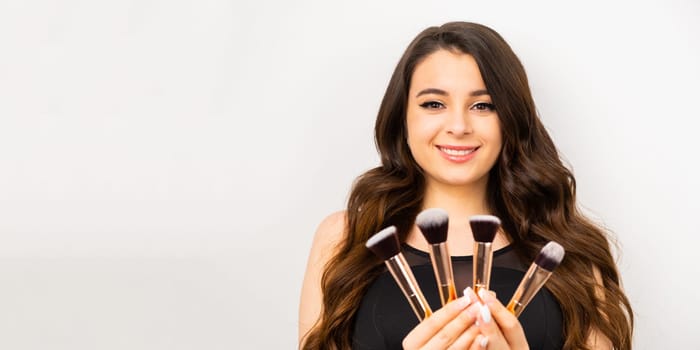 A smiling brunette woman holding makeup brushes on the white background.