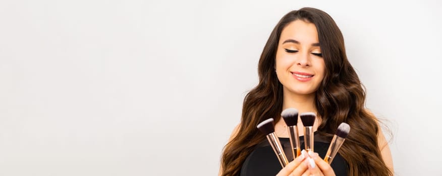 Young woman with makeup holding powder brush against grey background.