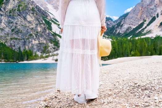 A young woman walks along the Lake Braies in a white dress and hat in her hands on the background of Dolomites