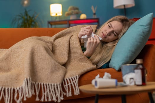 Young ill woman suffering from cold or allergy lying on sofa in living room at home apartment. Sick child girl with towel on head blows sneezes wipes snot into napkin. Coronavirus quarantine pandemic