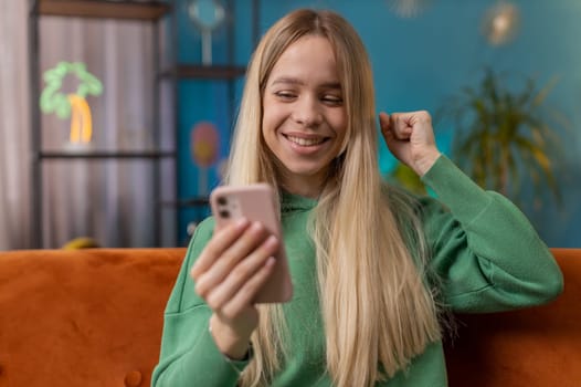 Happy woman use mobile smartphone typing browsing say Wow yes found out great big win good news celebrate lottery jackpot doing winner gesture. Teen young girl at home in room sitting on orange sofa