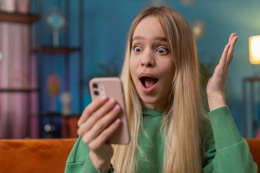 Oh my God Wow. Excited happy young woman use smartphone typing browsing celebrating success winning, lottery jackpot goal achievemen, good news, victory. Caucasian girl holds mobile phone at home room
