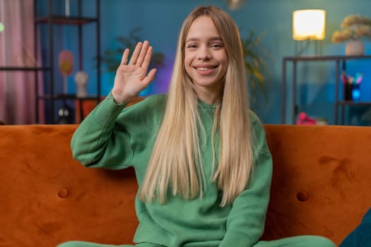 Hello. Caucasian woman smiling friendly at camera and waving hands gesturing hello, hi, greeting or goodbye, welcoming with hospitable expression at home. Teen girl sitting on sofa in living room