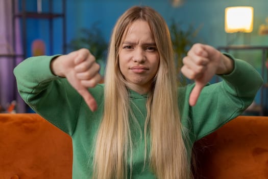Dislike. Upset young woman showing thumbs down sign gesture, expressing discontent, disapproval dissatisfied bad work at modern home apartment indoors. Displeased Caucasian girl in living room on sofa