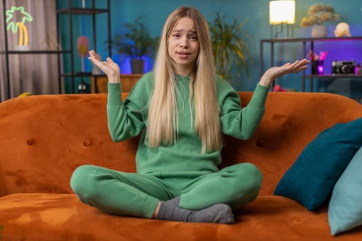 What. Why. Caucasian woman raising hands in indignant expression asking reason of failure demonstrating disbelief irritation by troubles at home apartment. Young girl in living room sitting on sofa