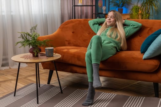 Tired woman lying down on comfortable couch, taking a rest, daytime nap. Portrait of carefree Caucasian girl holding hands behind head relaxing, falling asleep at home apartment living room on sofa