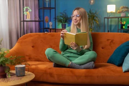 Young woman relaxing interesting book, turning pages smiling enjoying literature, taking a rest on comfortable couch. Portrait of peaceful cheerful Caucasian girl at home apartment living room on sofa