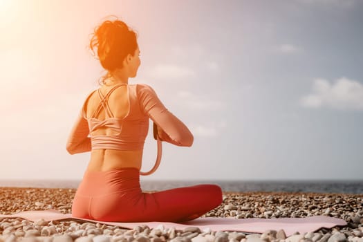 Middle aged well looking woman with black hair doing Pilates with the ring on the yoga mat near the sea on the pebble beach. Female fitness yoga concept. Healthy lifestyle, harmony and meditation.