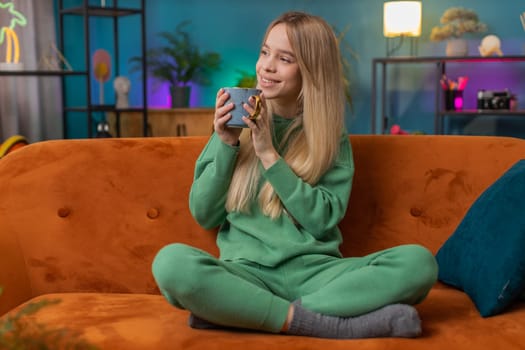 Portrait of smiling woman drinking a cup of warm coffee or herbal tea in the morning. Caucasian teen girl enjoying comfortable relaxing taking a break alone at home apartment living room on sofa