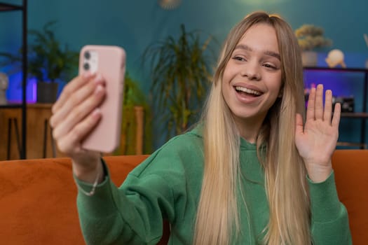 Happy woman blogger taking selfie on smartphone, communicating video call online with subscribers, recording stories for social media. Portrait of Caucasian girl at home apartment living room on sofa