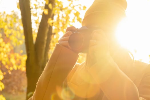 A woman photographer in a brown coat takes a photo on a retro camera in an autumn forest against sunlight, side view.