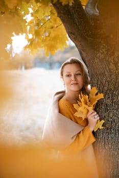 The blonde stands near the autumn tree. Thoughtful woman looks ahead, dressed in a yellow dress. Autumn content.