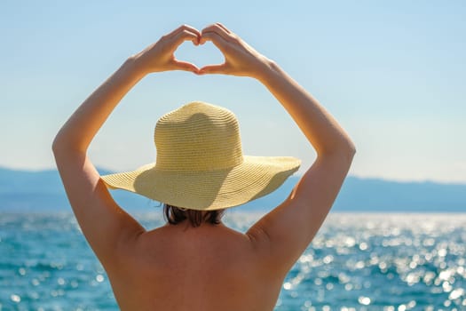 Lady in straw hat stands against blue sea showing heart shaped hands over head. Young woman looks at sparkling sea surface admiring nature beauty