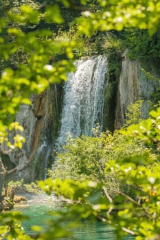 Waterfall flows down from rocky cliff into clear turquoise water at sunlight. Breathtaking view of aquamarine cascade in national park on Plitvice lakes