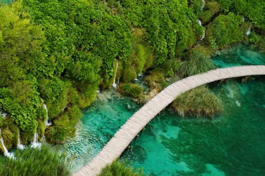 Hiking tourists walk on wooden footbridge surrounding lake with turquoise water in natural reserve. Majestic view of blue lake among picturesque landscape on Plitvice lakes upper view