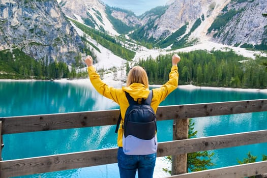 Tourist in yellow jacket and with backpack holds up her hands and enjoying the view of Lake Braies with turquoise water and Dolomites.