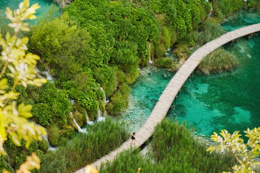 Hiking tourists walk on wooden footbridge surrounding lake with turquoise water in natural reserve. Majestic view of blue lake among picturesque landscape on Plitvice lakes upper view