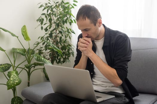 Confused man sitting on couch with computer on lap in living room, upset frustrated male reading bad news on message, having problem with device or Internet connection.