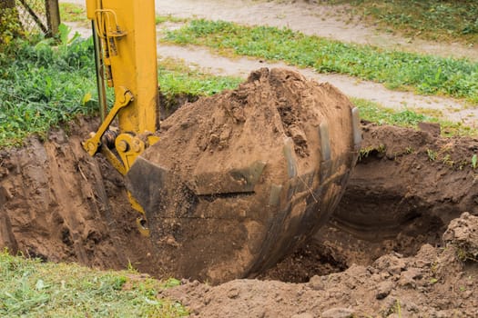 Excavator bucket with a pile of earth digs a large deep hole in the industrial area of the construction site.