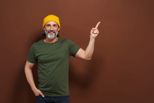 Smiling European man in a yellow hat points his hand to the side with copy space on a brown background. Product placing, advertising concept. High quality photo