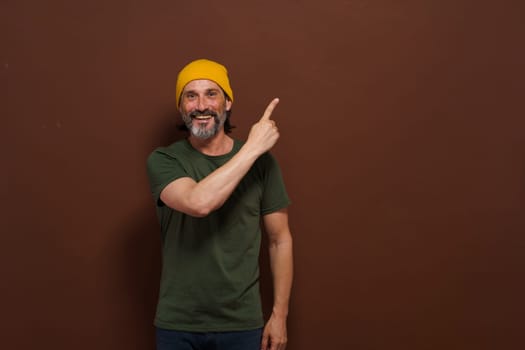Excited smiling Caucasian man in a yellow hat points his hand to the side with copy space on a brown background. Product placing, advertising concept. High quality photo