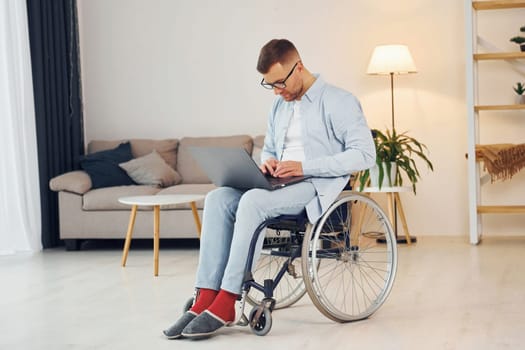 With laptop on the legs. Disabled man in wheelchair is at home.