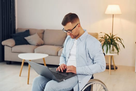 With laptop on the legs. Disabled man in wheelchair is at home.
