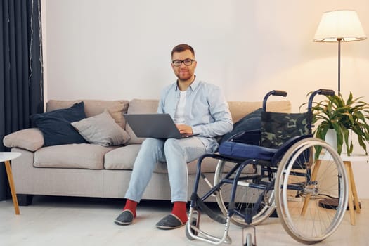 With laptop on the legs. Disabled man in wheelchair is at home.