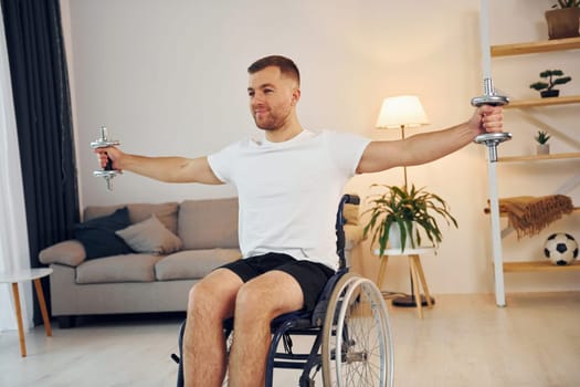 Lifting dumbbells. Disabled man in wheelchair is at home.