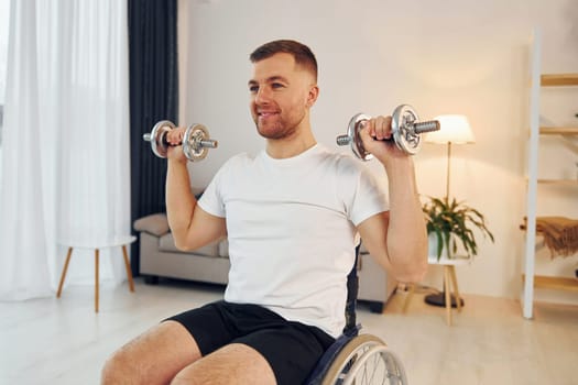 Doing exercises by using dumbbells. Disabled man in wheelchair is at home.