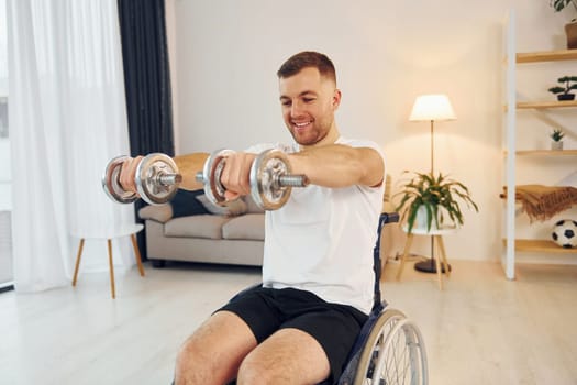 Holding weights. Disabled man in wheelchair is at home.