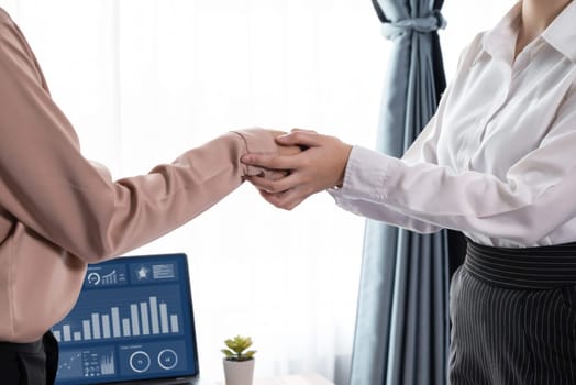 Two Asian businesswomen shaking hands in modern office after successfully analyzing piles of dashboard data papers, as importance of teamwork and integrity in the workplace concept. Enthusiastic