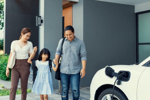Progressive young parents and daughter with electric vehicle and home charging station. Green and clean energy from electric vehicles for healthy environment. Eco power from renewable source at home.
