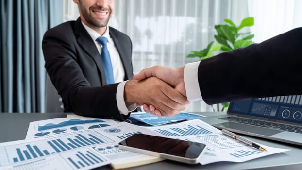 Closeup professional businessman shaking hands over desk in modern office after successfully analyzing pile of dashboard data paper as teamwork and integrity handshake in workplace concept. fervent