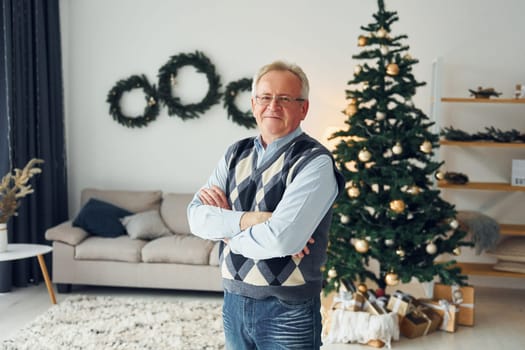 Celebrating new year and standing near christmas tree. Senior man in nice clothes is at home.