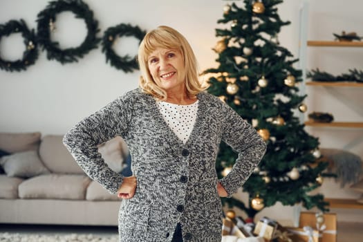 Standing near christmas tree. Senior woman with blonde hair is at home.