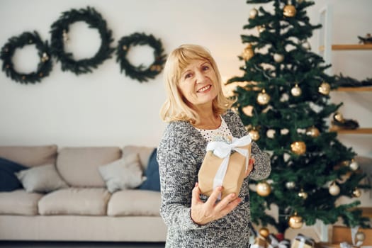 Standing near christmas tree. Senior woman with blonde hair is at home.