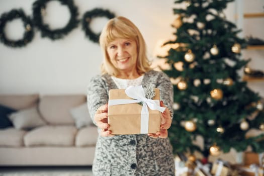 Standing near christmas tree. Senior woman with blonde hair is at home.