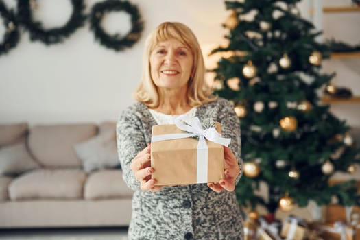 Standing near christmas tree. Senior woman with blonde hair is at home.