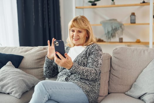 Sitting and holding phone. Senior woman with blonde hair is at home.