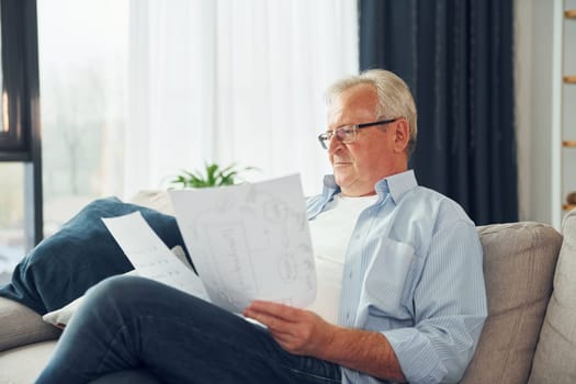 Holding two papers. Senior man in nice clothes is at home.