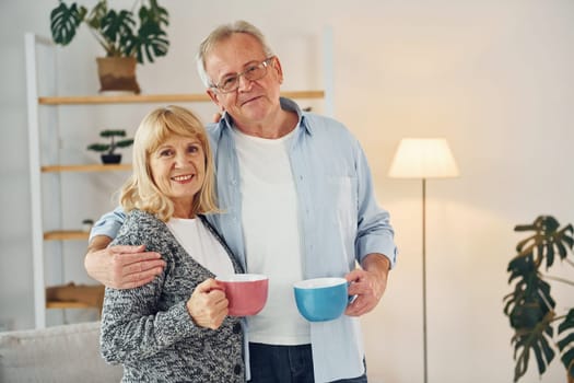 Standing and holding cups of drinks. Senior man and woman is together at home.