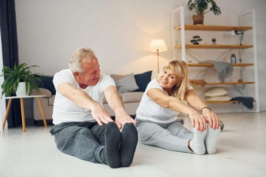 Sitting and doing exercises. Senior man and woman is together at home.