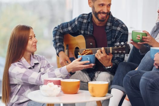 Man is playing acoustic guitar. Group of friends have party indoors together.