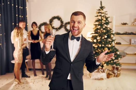 Man with glass of champagne is cheering. Group of people have a new year party indoors together.