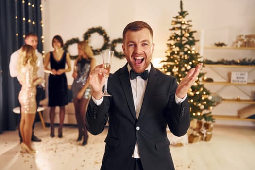 Man with glass of champagne is cheering. Group of people have a new year party indoors together.