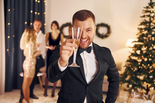 Man with glass of champagne is cheering. Group of people have a new year party indoors together.